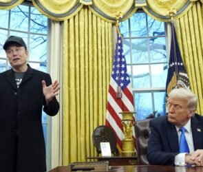 Elon Musk stands in an Office, gesturing, while Donald Trump, the US President sits at the Resolute Desk, looking at him. The American flag stands behind them.