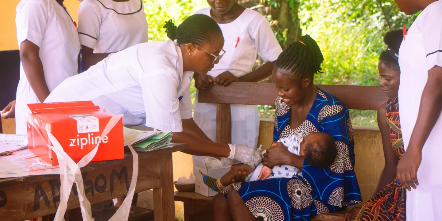 A Zipline medical delivery box sits on the table while a nurse administers a vaccine to a baby held by a smiling mother at a rural clinic, with other nurses observing in the background.