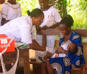 A Zipline medical delivery box sits on the table while a nurse administers a vaccine to a baby held by a smiling mother at a rural clinic, with other nurses observing in the background.