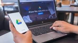 An human being holding a smartphone displaying Google Drive icon alongside a laptop